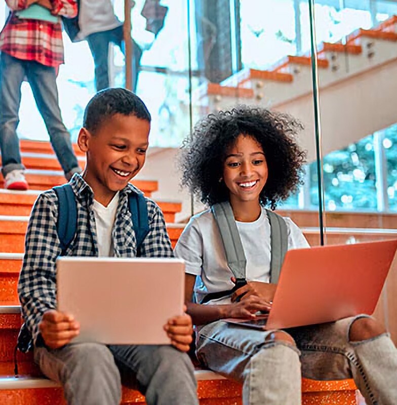 Two children smiling while using laptops while seated in the school staircase
